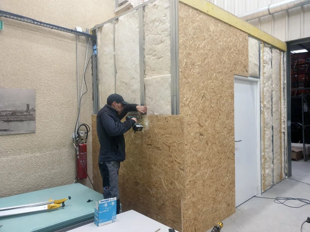 Portrait en situation d'un artisan-entrepreneur dans un intérieur en cours de rénovation, discutant attentivement avec un propriétaire. L'artisan consulte un plan ou un document détaillé sur un support mobile ou papier, écoutant activement le client avec une expression concentrée et professionnelle. En arrière-plan, des éléments de rénovation visibles (outils, matériaux de qualité, espace partiellement rénové) illustrant le contexte du métier. La scène se déroule dans un vrai espace résidentiel en rénovation, lumière naturelle, sans mise en scène forcée. L'accent visuel doit être sur l'échange conseil authentique entre l'artisan expert et le client, reflétant l'approche "conseil avant devis" de l'entreprise.