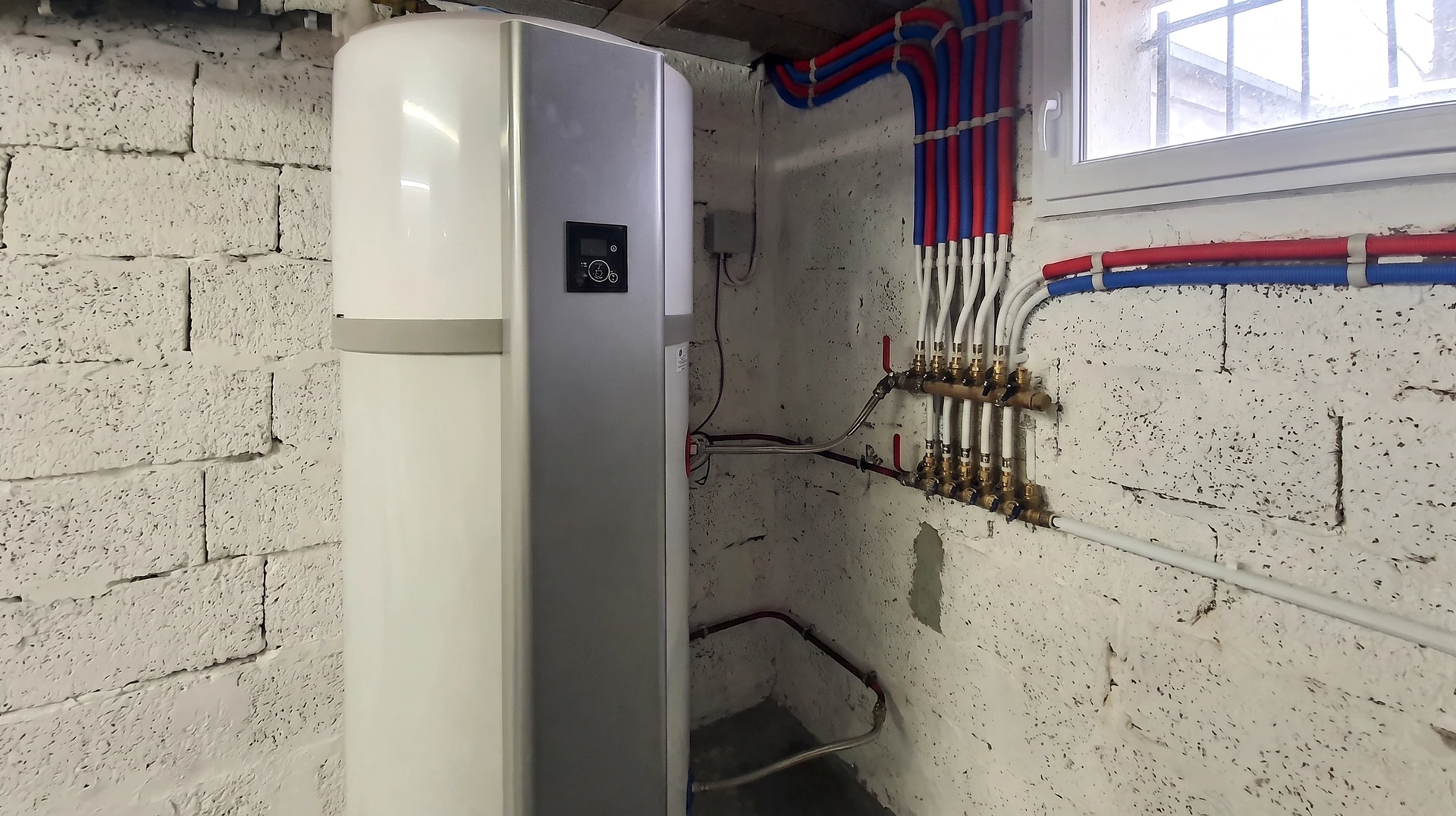 Photo en gros plan d'un plombier expert en train d'installer un ballon d'eau chaude thermodynamique ou électrique moderne dans un intérieur résidentiel en cours de rénovation. L'artisan ajuste les raccordements cuivre ou flexibles, concentré sur son travail. On voit clairement le chauffe-eau blanc ou noir, les tuyauteries bien organisées, et l'environnement de rénovation (murs nus ou partiellement finis). La scène capture professionnalisme, expertise et fiabilité technique. Lumière naturelle mettant en avant la qualité de l'exécution.